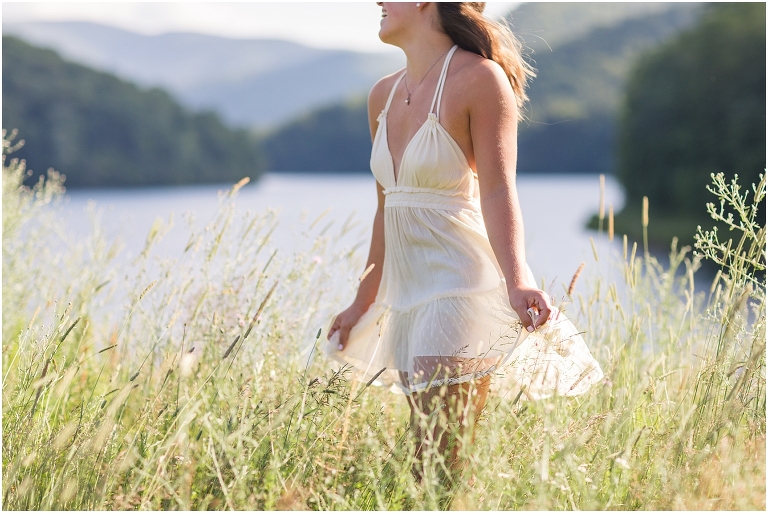 Senior photos of a girl in a white dress in a field by the lake with a mountain view taken by a Virginia graduation photographer