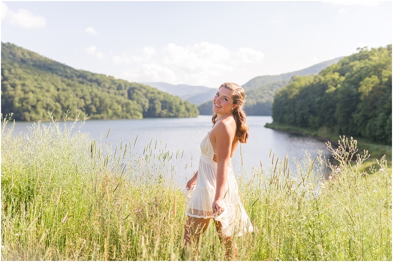 Senior photos of a girl in a white dress in a field by the lake with a mountain view taken by a Virginia graduation photographer