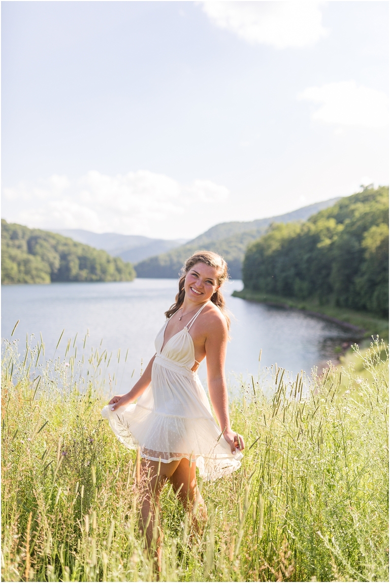 Senior photos of a girl in a white dress in a field by the lake with a mountain view taken by a Virginia graduation photographer