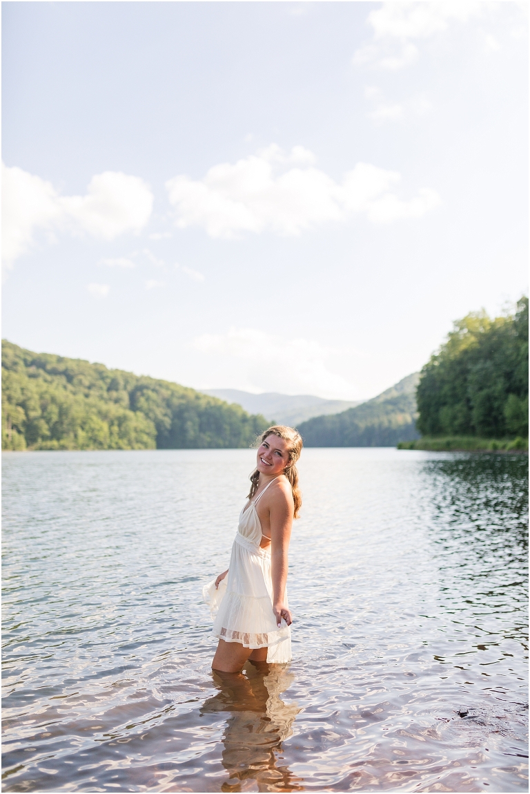 Senior photos of a girl in a white dress in a field by Switzer Lake with a mountain view taken by a Virginia graduation photographer