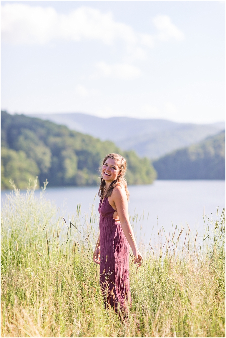 Senior photos of a girl in a purple dress in a field overlooking a lake with a mountain background taken by a Virginia portrait photographer