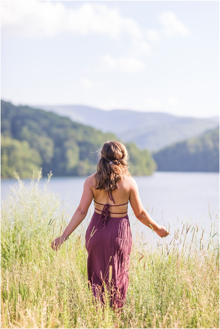 Senior photos of a girl in a purple dress in a field overlooking a lake with a mountain background taken by a Virginia portrait photographer