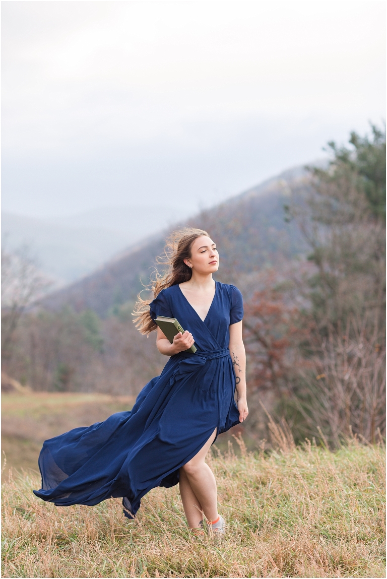 Windy senior portraits in a blue dress holding a book next to Switzer Lake with a mountain background at sunset, Virginia senior graduation portrait photographer